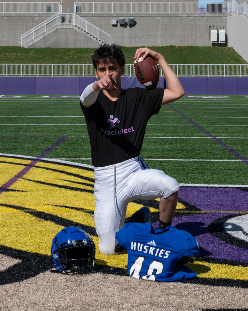 A teen boy who was born with clubfoot wears a MiracleFeet shirt while posing on a football field.